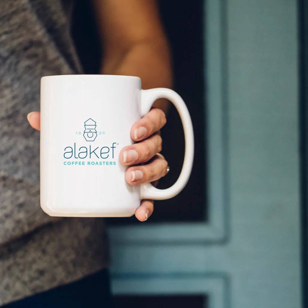 person holding white ceramic mug with alakef coffee roasters logo and branding., alakef coffee roasters inc, duluth