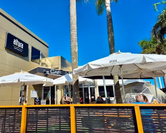 beachside café with yellow fencing, white umbrellas, and tall palm trees against blue sky., altura coffee company, auckland