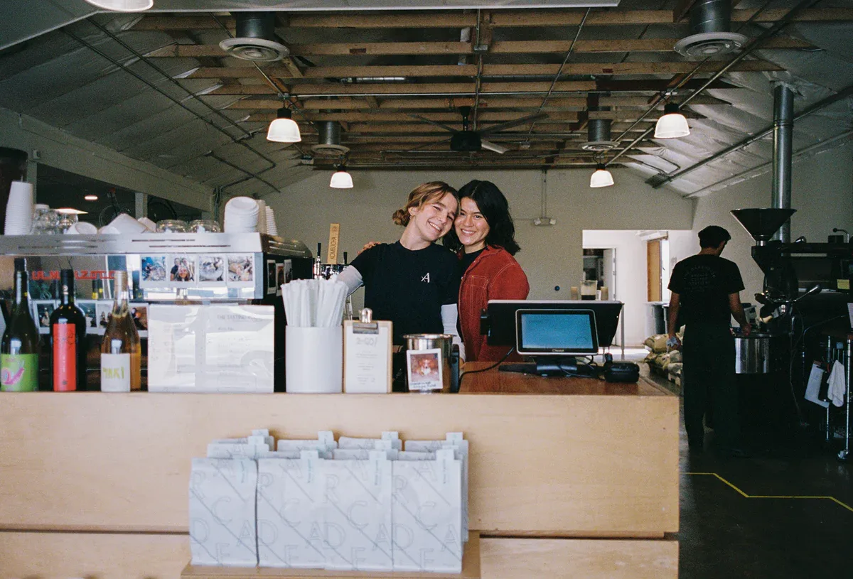 two smiling staff members behind a coffee shop counter with laptop and supplies., arcade coffee roasters - tasting room, rive