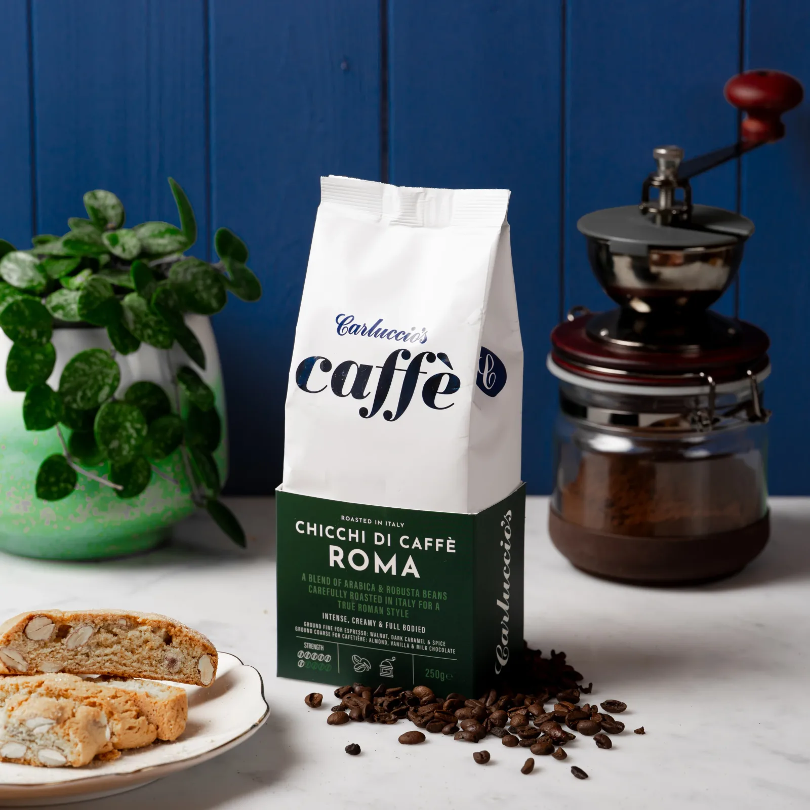 carluccio's caffè roma coffee bag displayed with grinder, biscotti, and potted plant against blue wall., artigiano del caffè,
