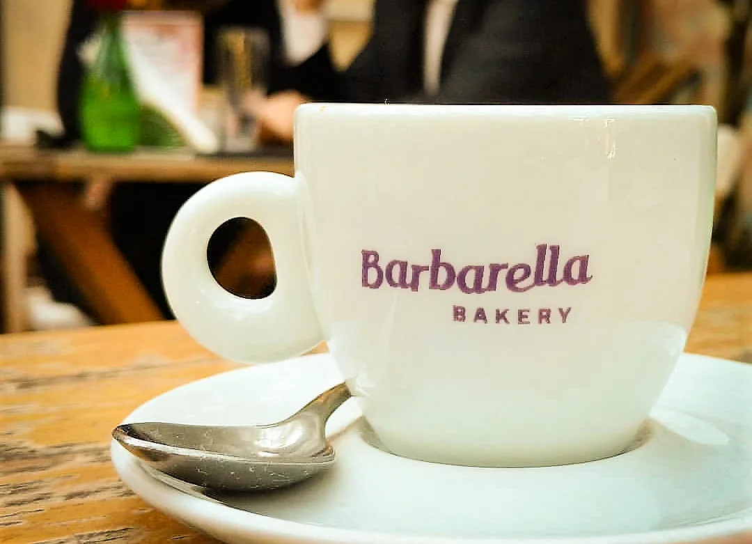 white mug with "barbarella bakery" text, saucer, spoon, wooden table, blurred background., barbarella bakery, curitiba