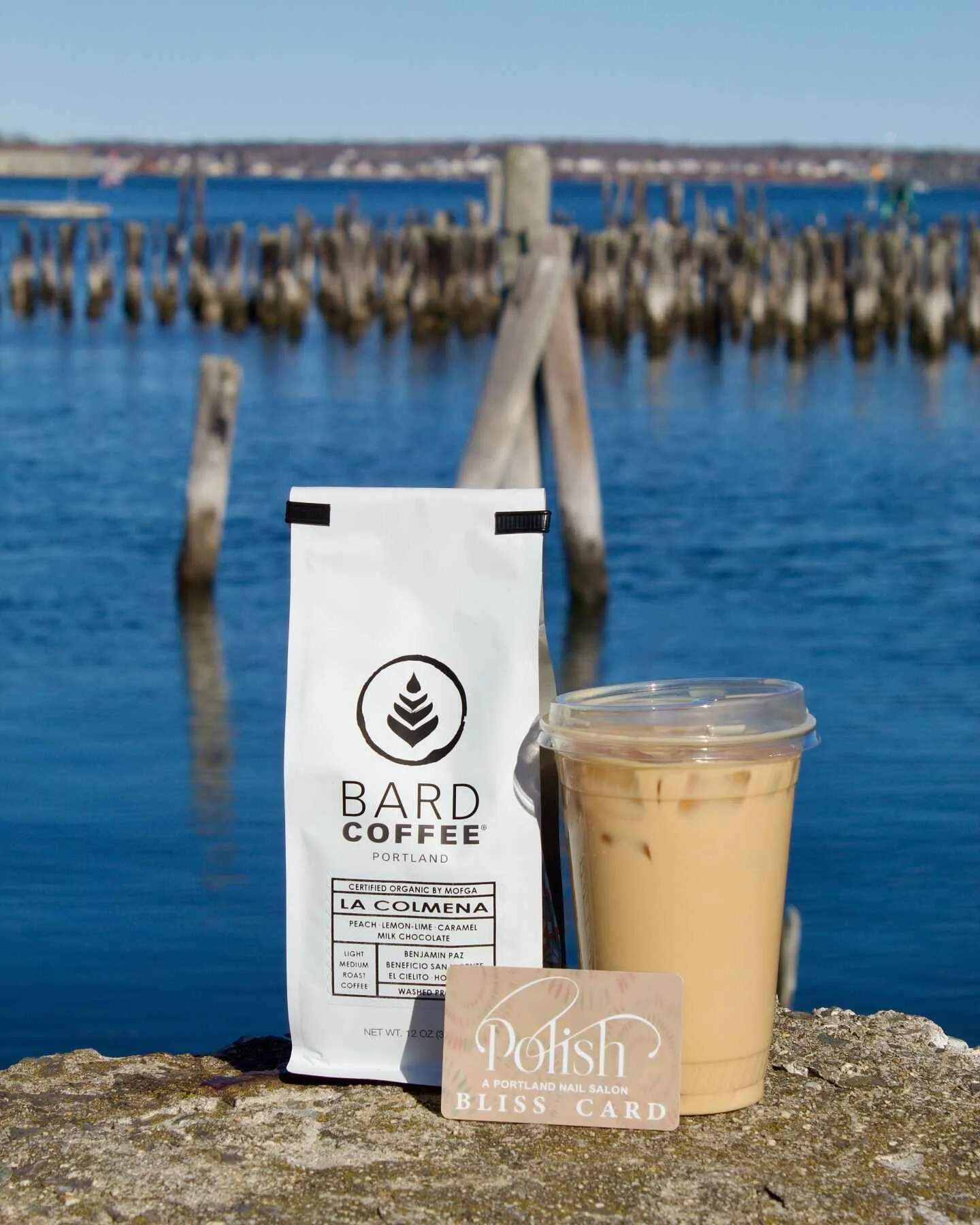 bard coffee bag and iced latte beside waterfront pier with wooden posts., bard coffee, portland
