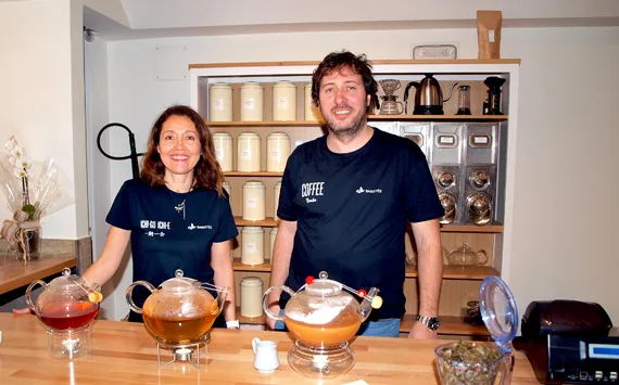 two people in matching navy t-shirts standing behind wooden counter with teapots and shelving., baristés, terrassa
