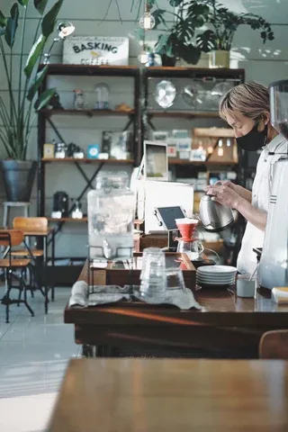 barista preparing drink at counter with shelves of plants and cafe supplies behind., basking coffee, fukuoka