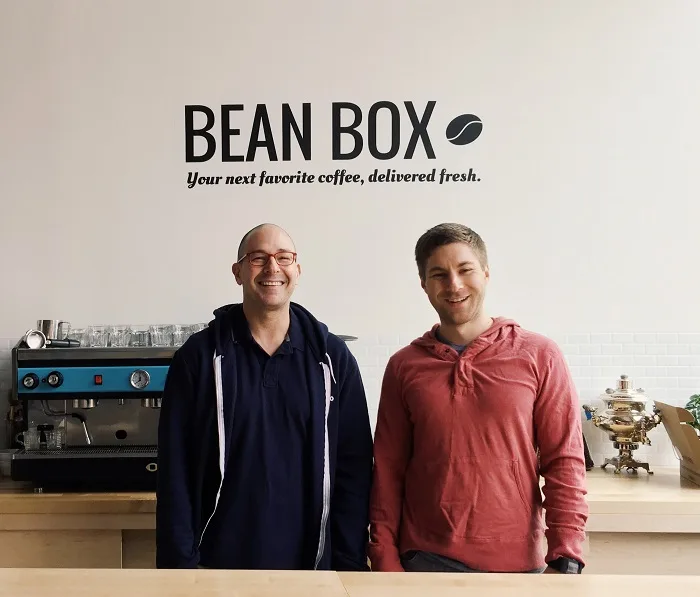 two men standing in front of coffee equipment and bean box wall signage., bean box, seattle