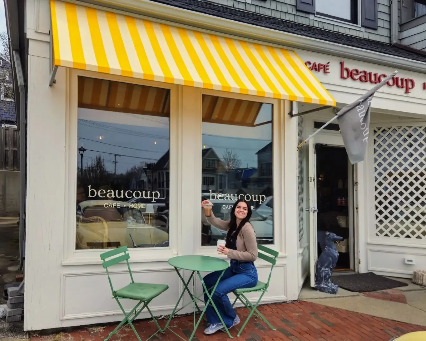 woman sits at green table outside yellow-striped café with coffee cup., beaucoup, newport