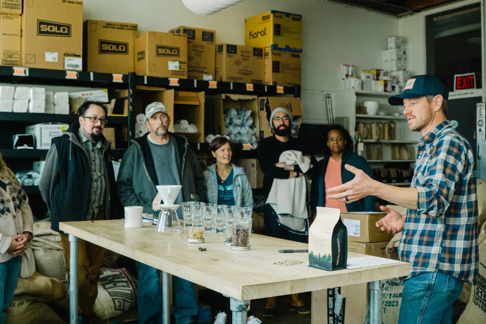 man in plaid shirt presents to group in warehouse with grain samples and boxes., building 3 coffee, colorado springs