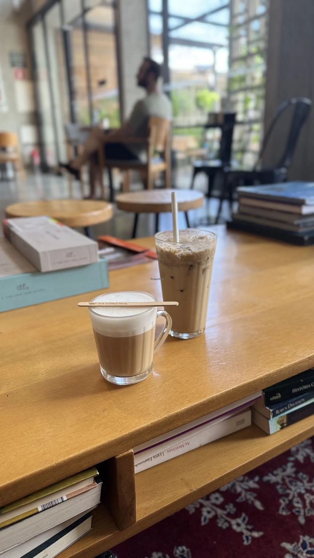 two coffee drinks on wooden table in bright, modern café with person in background., café cultura, curitiba