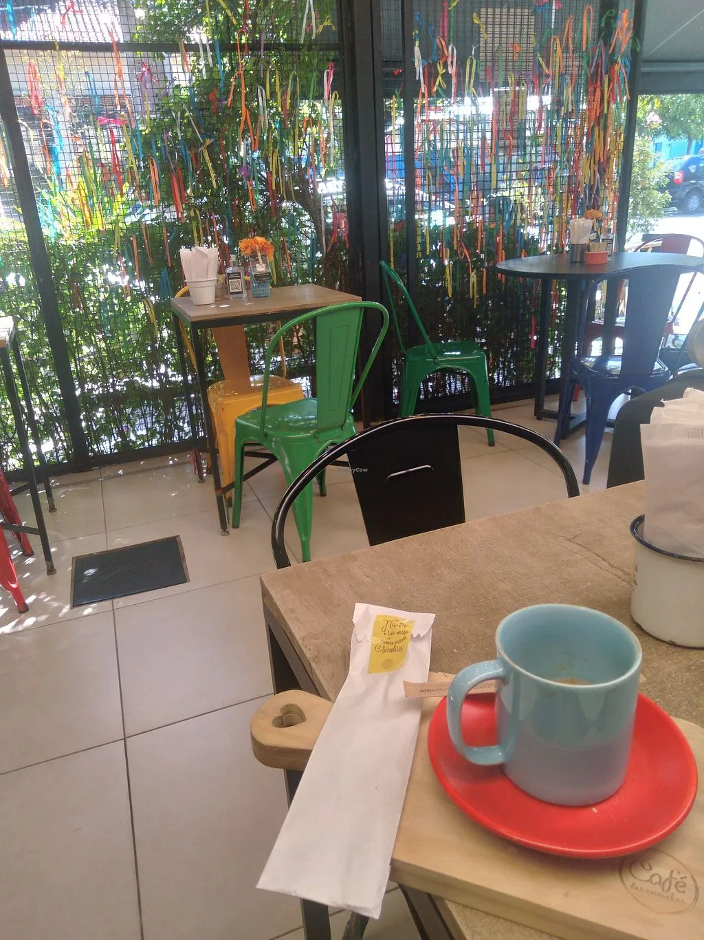 café interior with colorful hanging decorations, green chair, blue mug on red saucer., café das coisinhas, são paulo