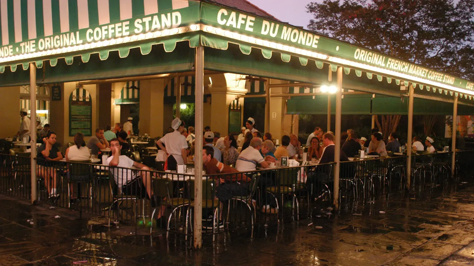 bustling outdoor café with green striped awning, wet pavement, evening patrons seated at tables., cafe du monde, new orleans