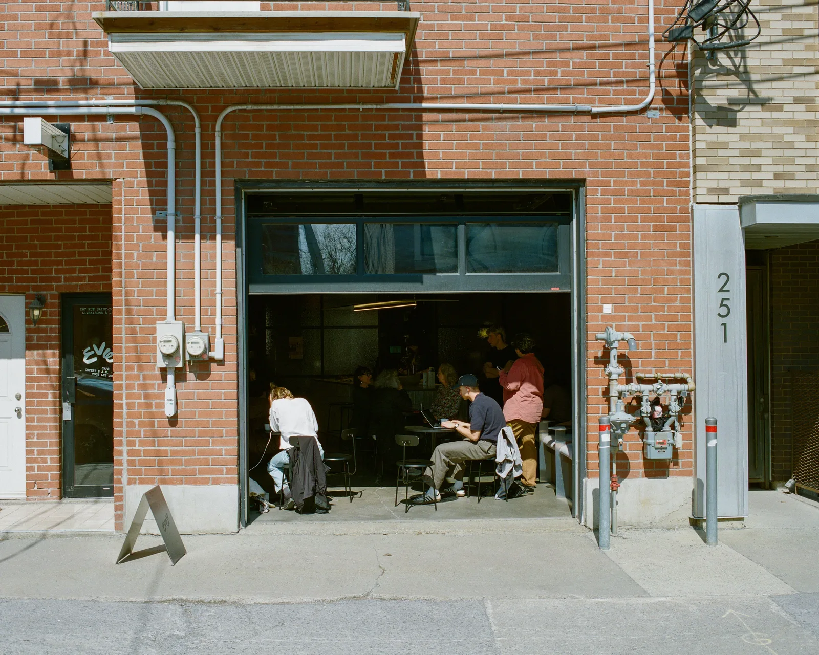 red brick storefront with open garage door, people sitting inside working or socializing., café even, montreal