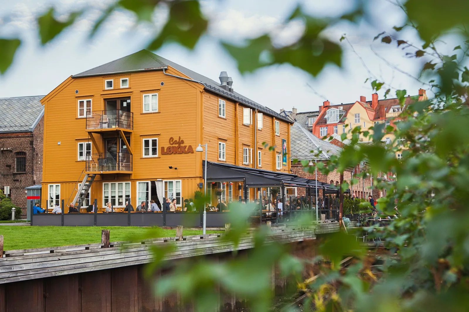 orange wooden building with café, riverside deck, historic architecture in background., café løkka, trondheim