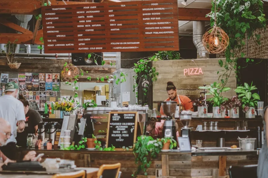 bustling café interior with wooden counter, pizza neon sign, and abundant greenery., cartel roasting co, tucson