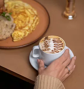 person holding cappuccino with latte art beside plate of eggs and toast., casa de tia empório & bistrô, curitiba