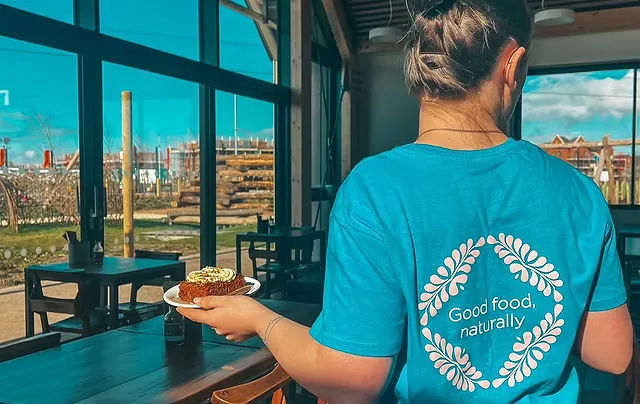 woman in blue "good food naturally" shirt holds pastry by large windows overlooking cityscape., cassiobury park, watford