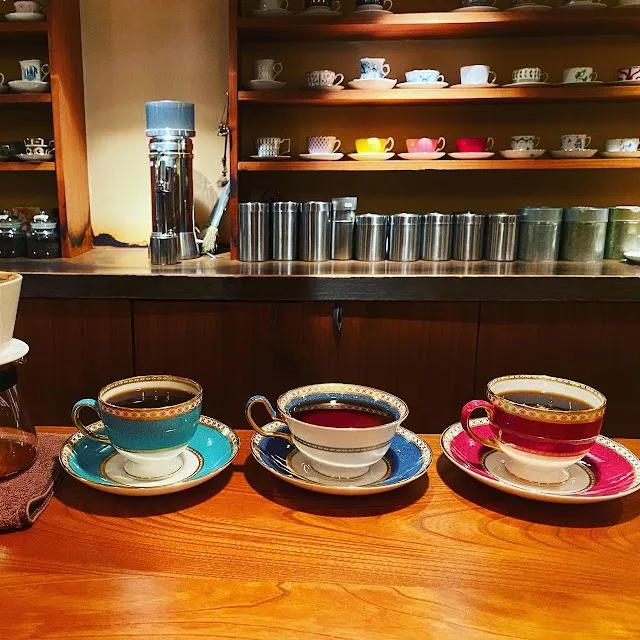 three elegant teacups with saucers displayed on wooden counter, shelves of ceramic dishware behind., charcoal roast coffee ri