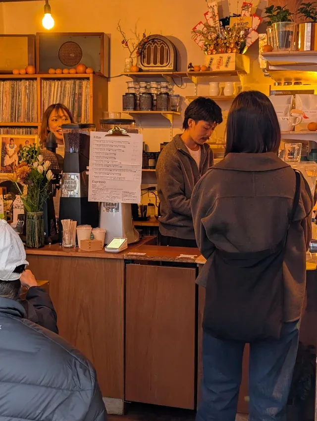 cozy café interior with warm lighting, customers at wooden counter, shelved décor and vintage radios., coffee swamp, tokyo