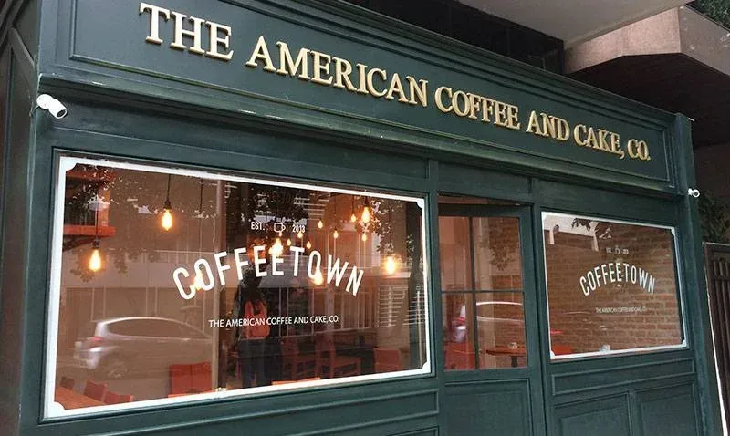 green storefront with "the american coffee and cake co" signage and warm interior lighting., coffeetown, rio de janeiro