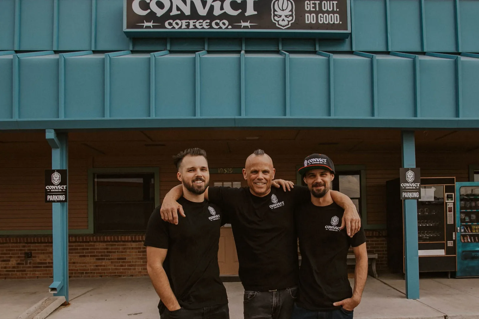 three men in matching black convict coffee co. shirts stand smiling outside storefront., convict coffee company, parker