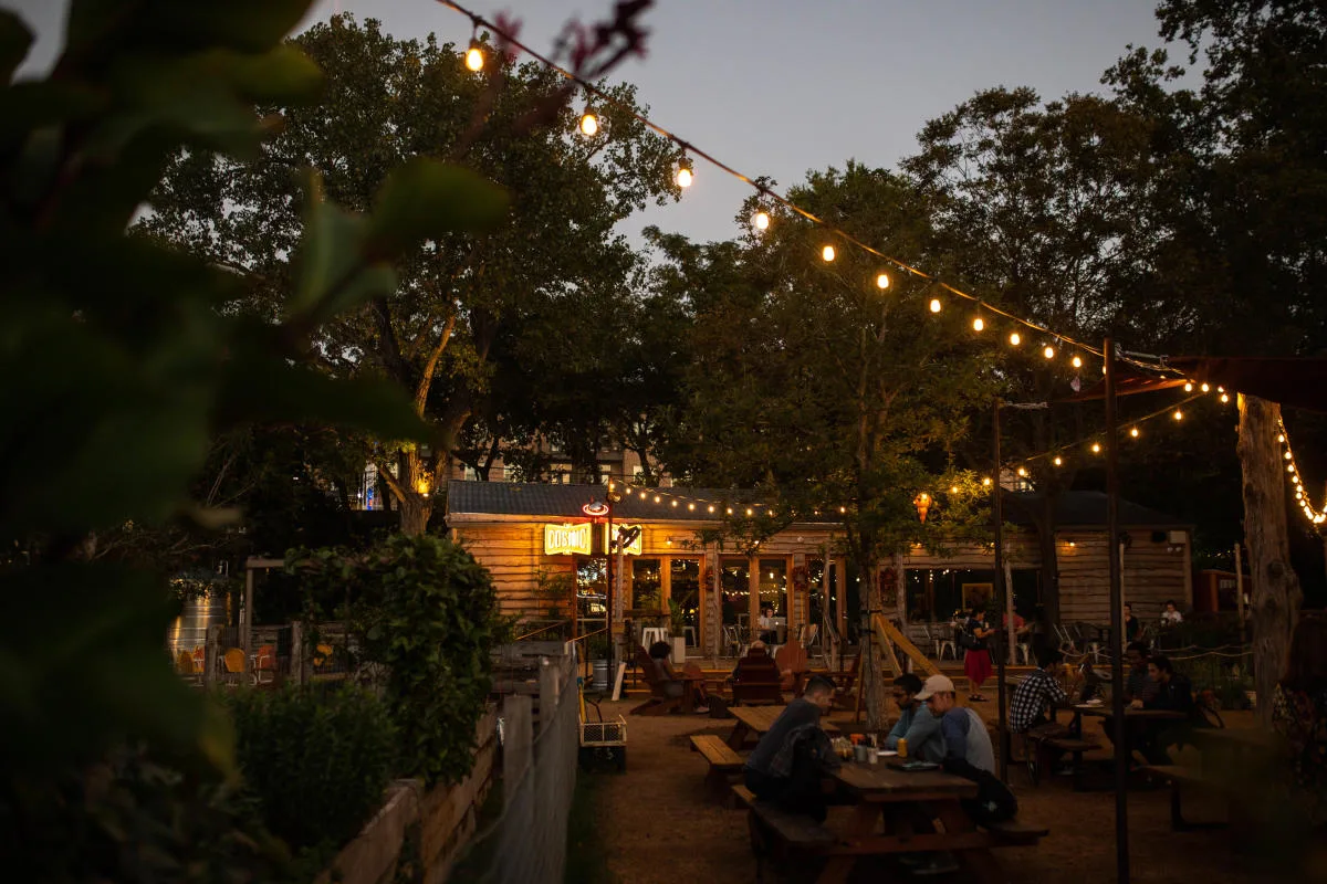 rustic outdoor venue with string lights, wooden building, and evening diners under trees., cosmic coffee + beer garden, austi