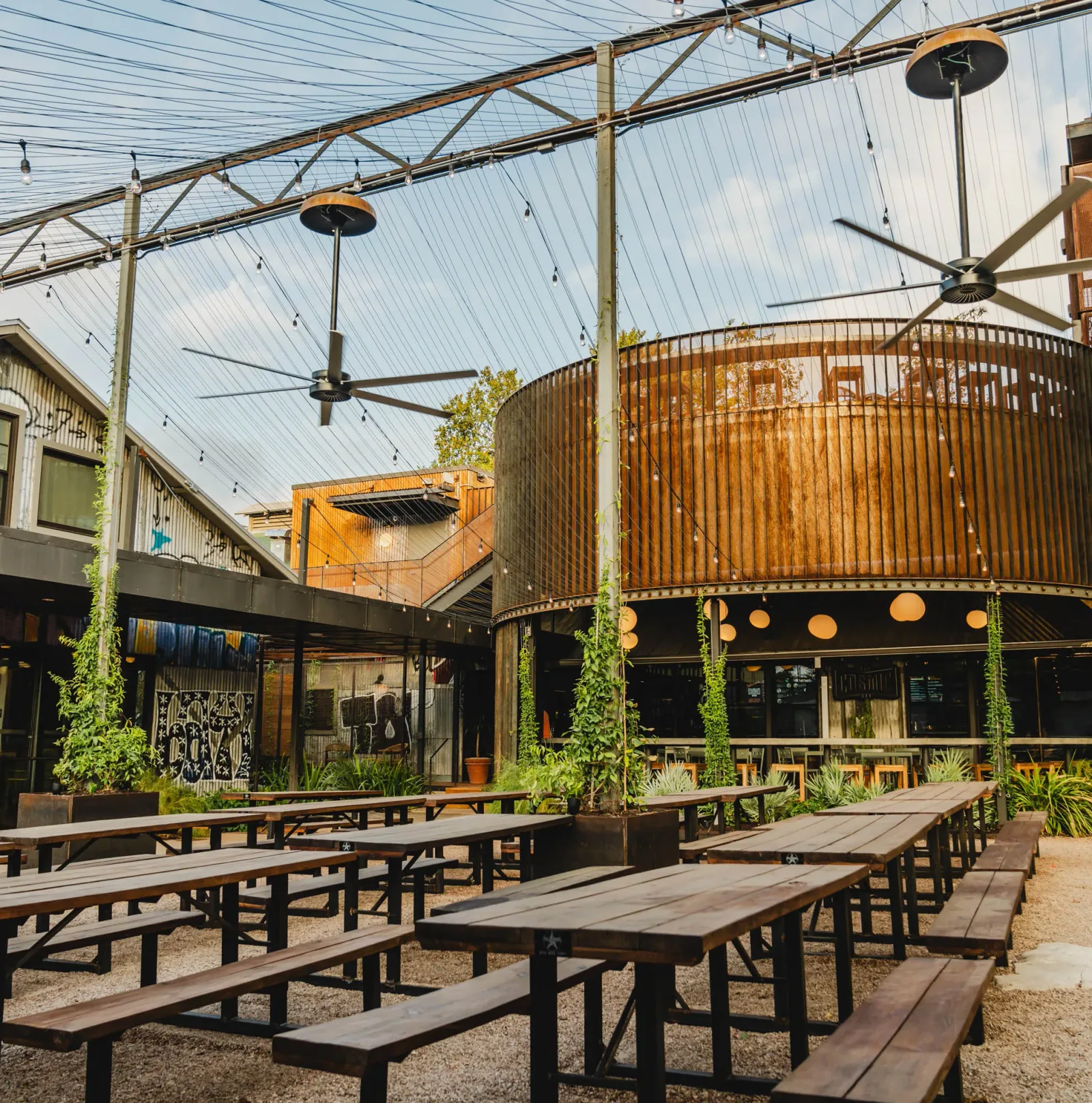 industrial outdoor patio with picnic tables, corrugated metal structures, and ceiling fans., cosmic coffee + beer garden, aus