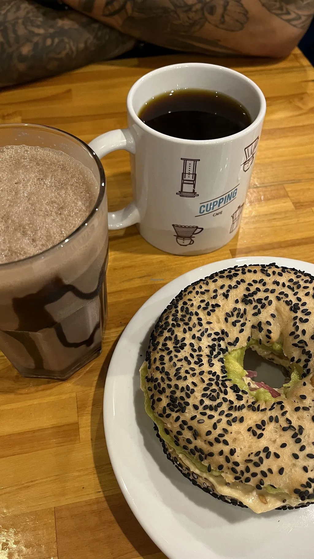 black sesame bagel with cream cheese, coffee mug, and beverage on wooden table., cupping café, são paulo