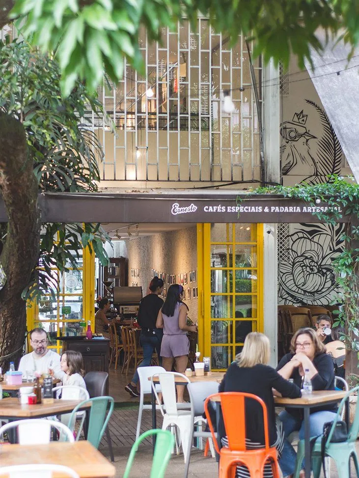 cozy café with yellow doors, outdoor seating, lush greenery, and customers inside., ernesto cafés asa norte, brasília