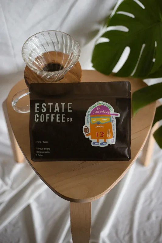 estate coffee co package with pour-over cup on wooden holder, monstera plant visible., estate coffee company, san antonio