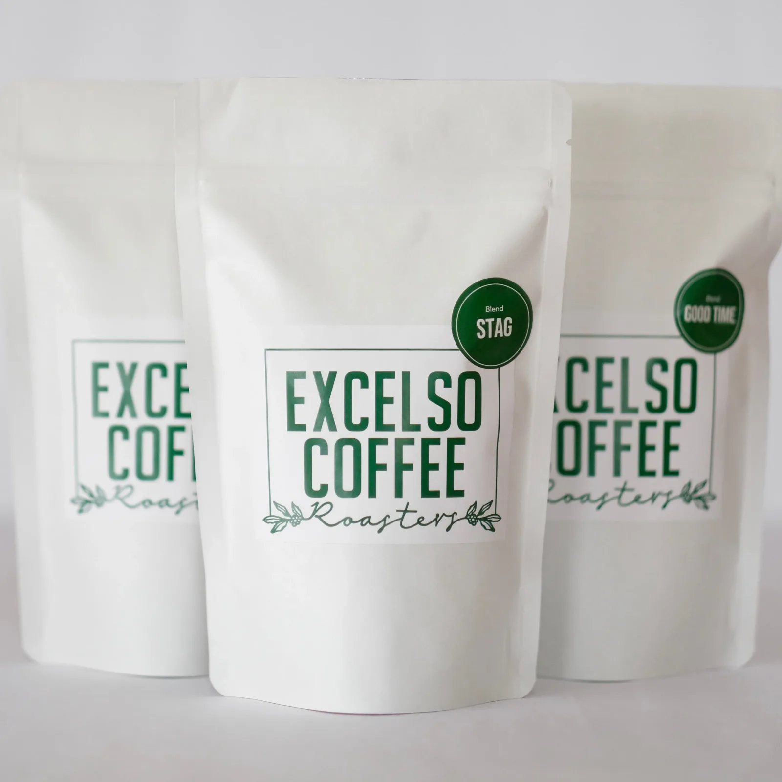 three white coffee pouches with green "excelso coffee roasters" branding and circular seal labels., excelso coffee, tauranga