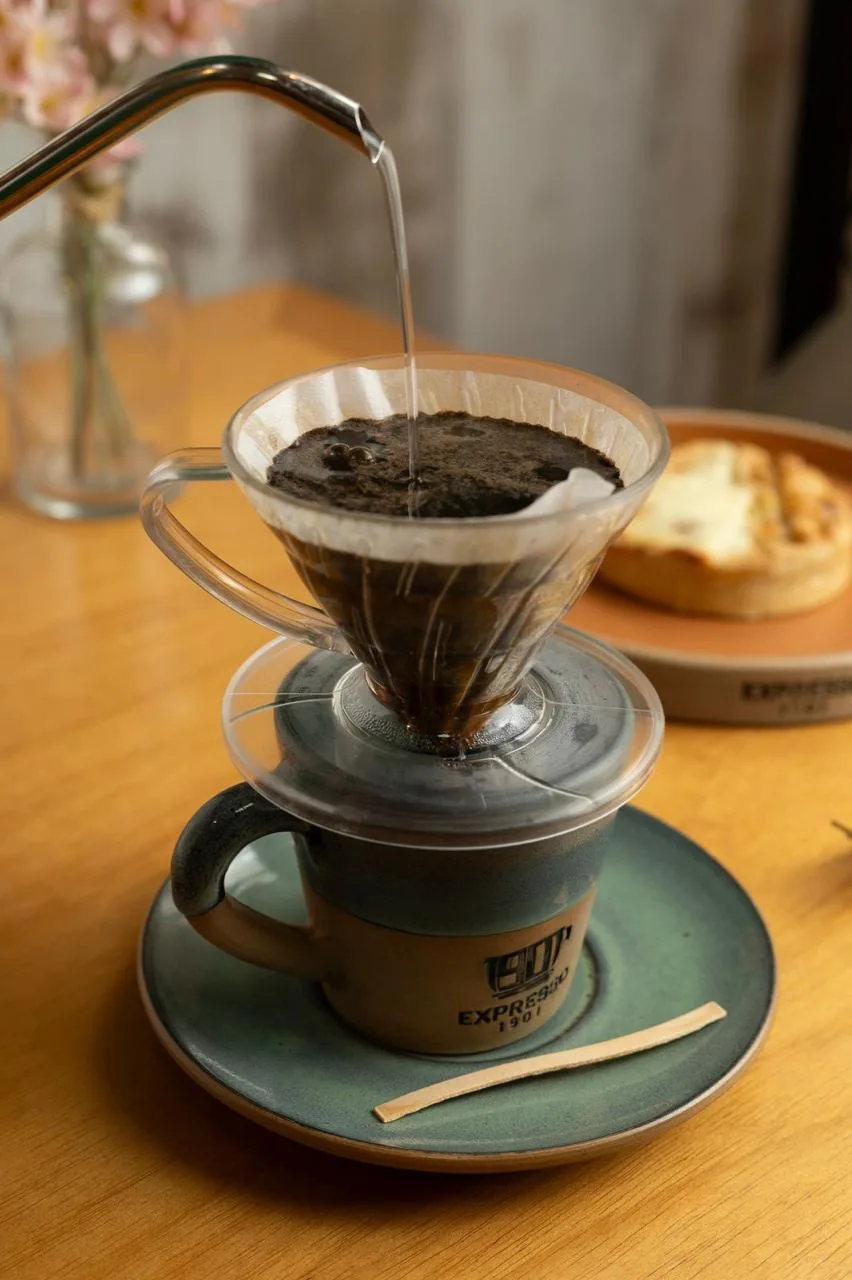 hot water pouring through coffee filter into ceramic cup with pastry nearby., expresso 1901, são paulo