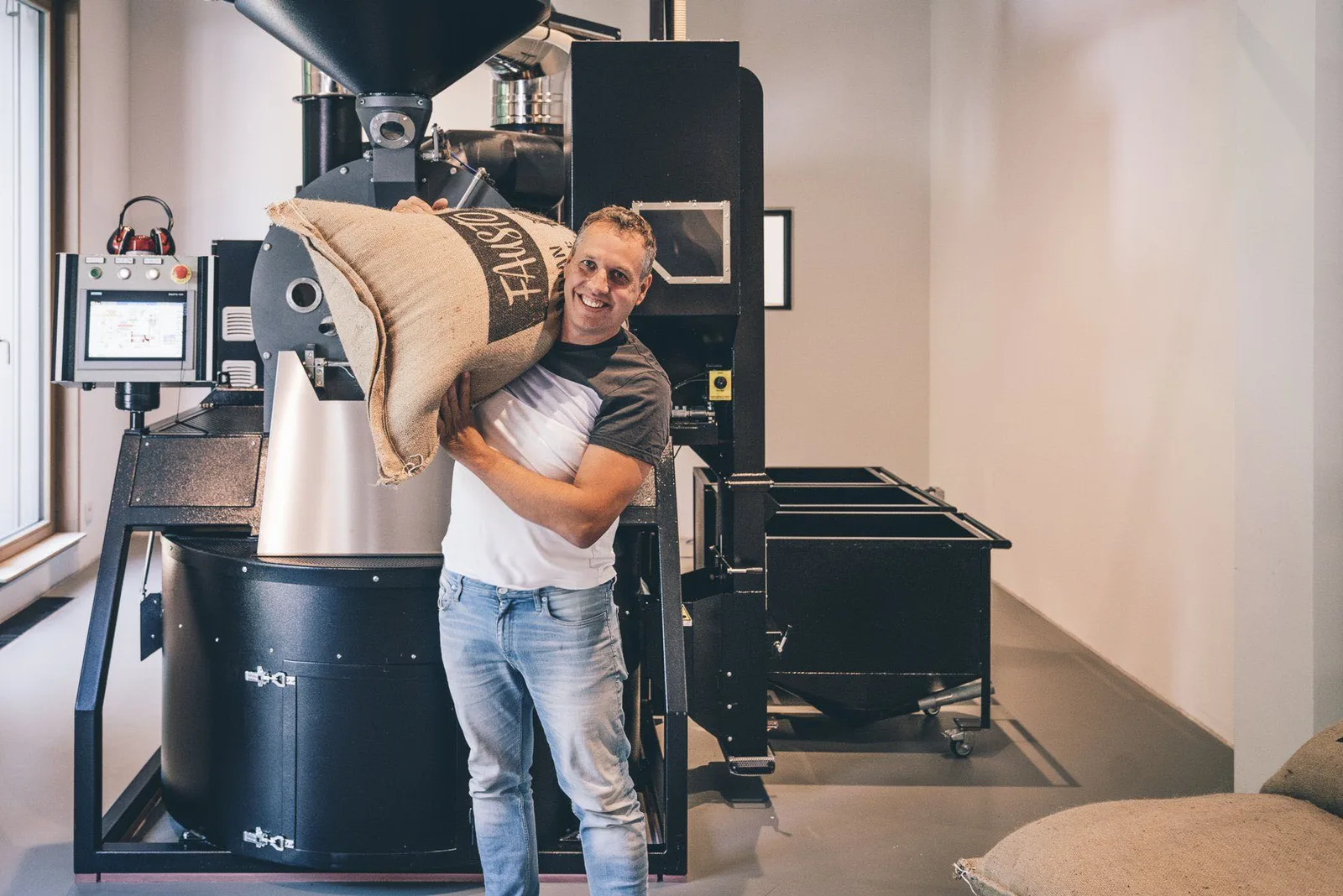 man holding burlap coffee sack beside industrial roasting machine., fausto kaffeerösterei gmbh, munich