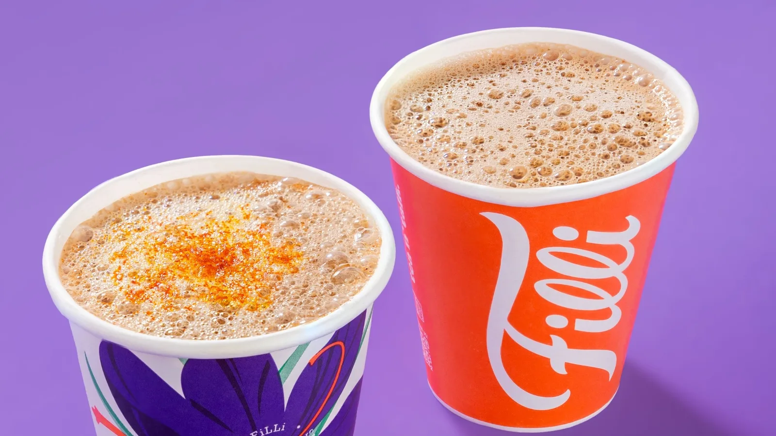 two cups of creamy coffee beverages on purple background, one with cinnamon topping., filli cafe, london