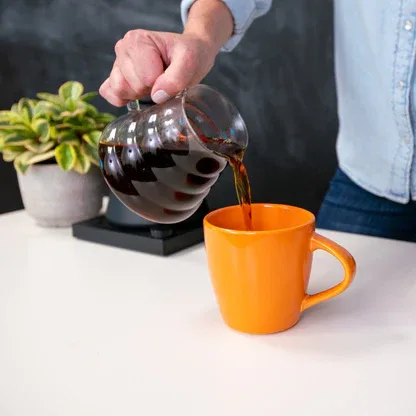 person pouring dark coffee from glass carafe into orange mug on white desk., fireside coffee co, flint