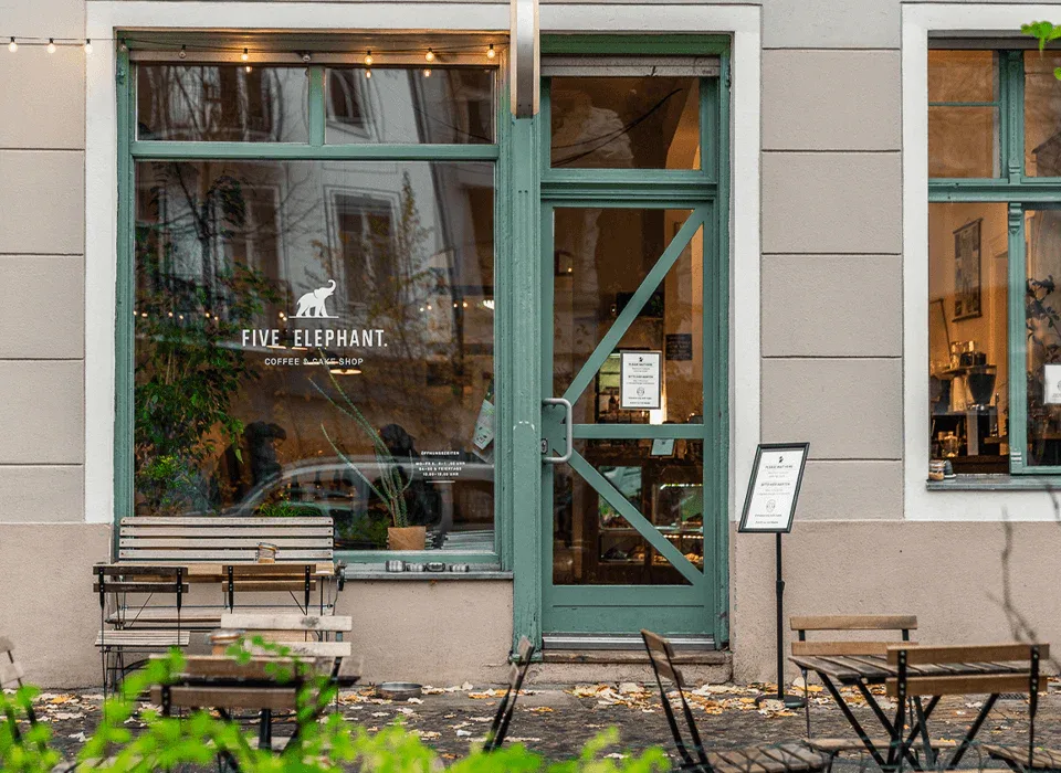 green-framed coffee shop storefront with benches and warm interior lighting., five elephant kreuzberg, berlin