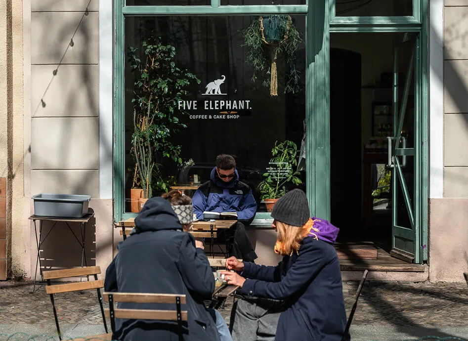 cozy coffee shop storefront with customers seated outdoors on wooden chairs., five elephant kreuzberg, berlin