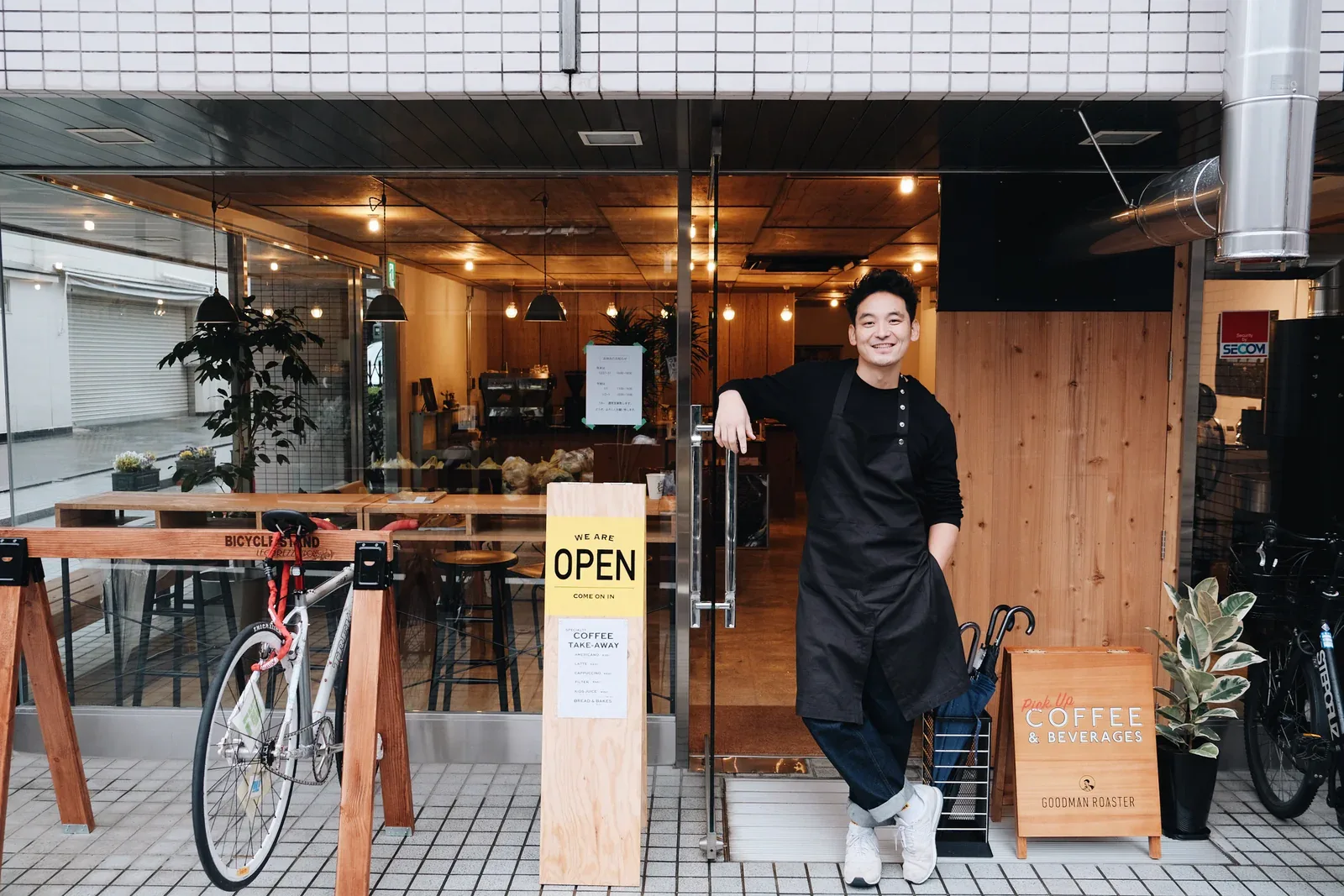 young man stands in doorway of warm-lit coffee shop with bicycles and wooden interior., goodman roaster, kyoto