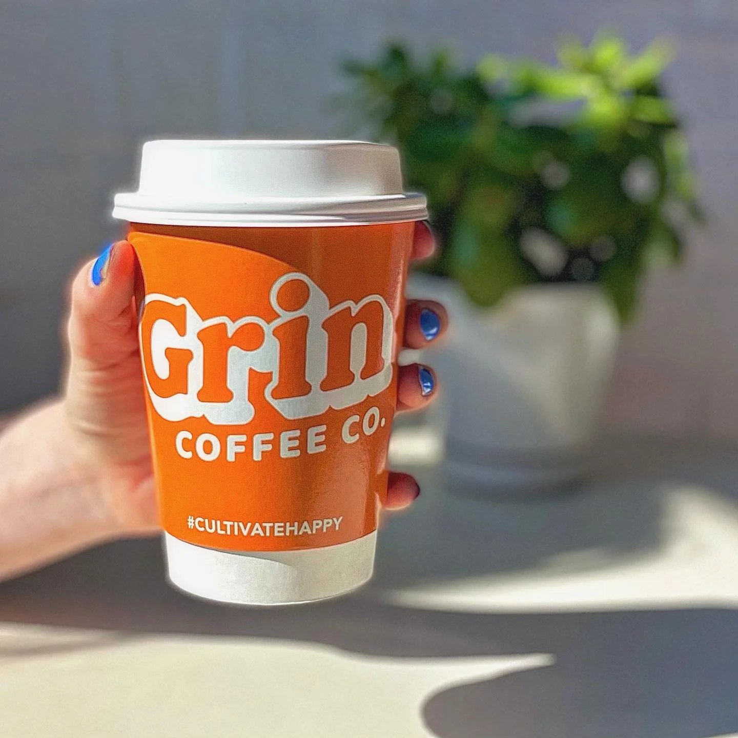 hand holding orange grin coffee co. cup with hashtag #cultivatehappy and potted plants behind., grin coffee roastery, mississ