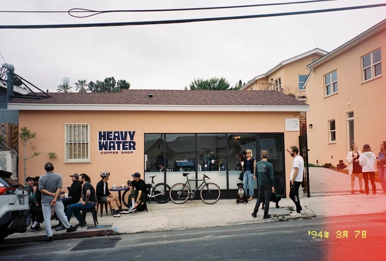 heavy water coffee shop storefront with customers, bicycles, and peach-colored buildings., heavy water coffee roasters, los a