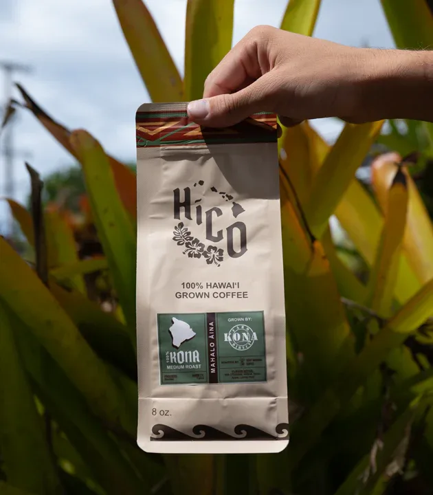 hand holding kona coffee package in front of green cactus plants outdoors., hico - hawaiian coffee, kona coffee, kailua-kona