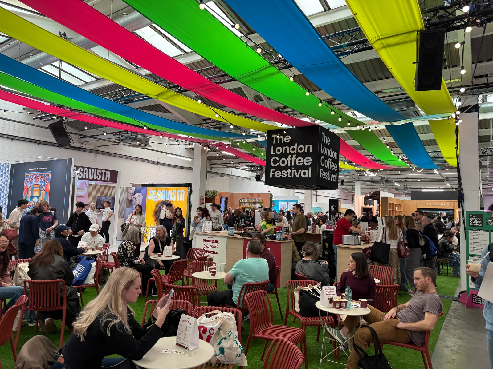 the london coffee festival main hall — colourful canopy drapes overhead, packed tables, lcf branded signage in the centre