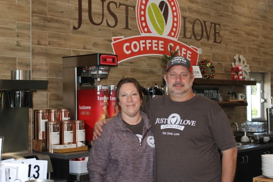 two people standing in front of "just love coffee cafe" counter with equipment and signage., just love coffee cafe - tomball,