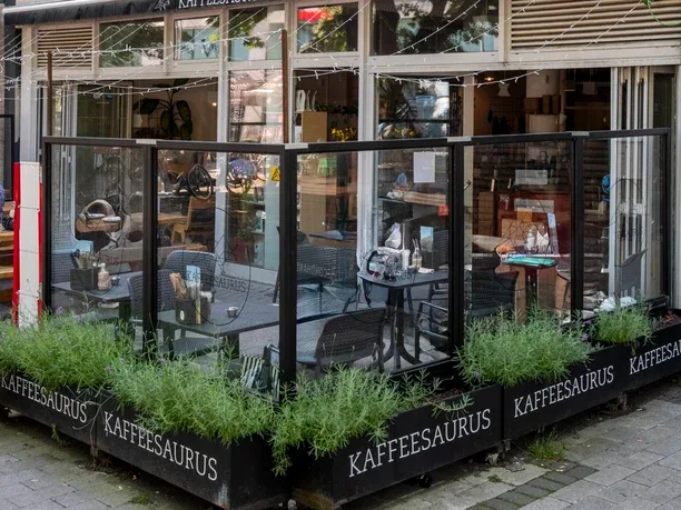 modern café exterior with glass frontage, black framing, and potted green plants., kaffeesaurus, cologne