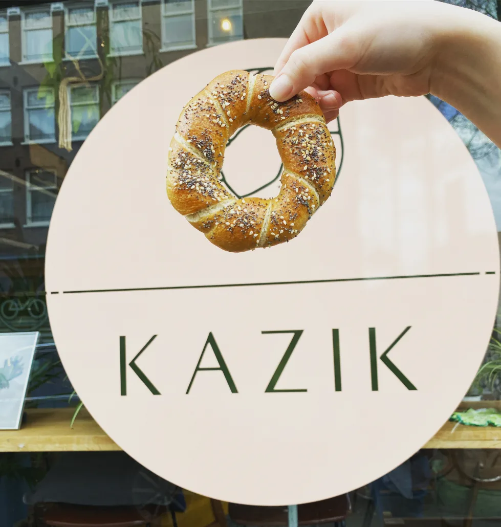 hand holding a sesame seed pretzel above kazik bakery sign., kazik amsterdam, amsterdam