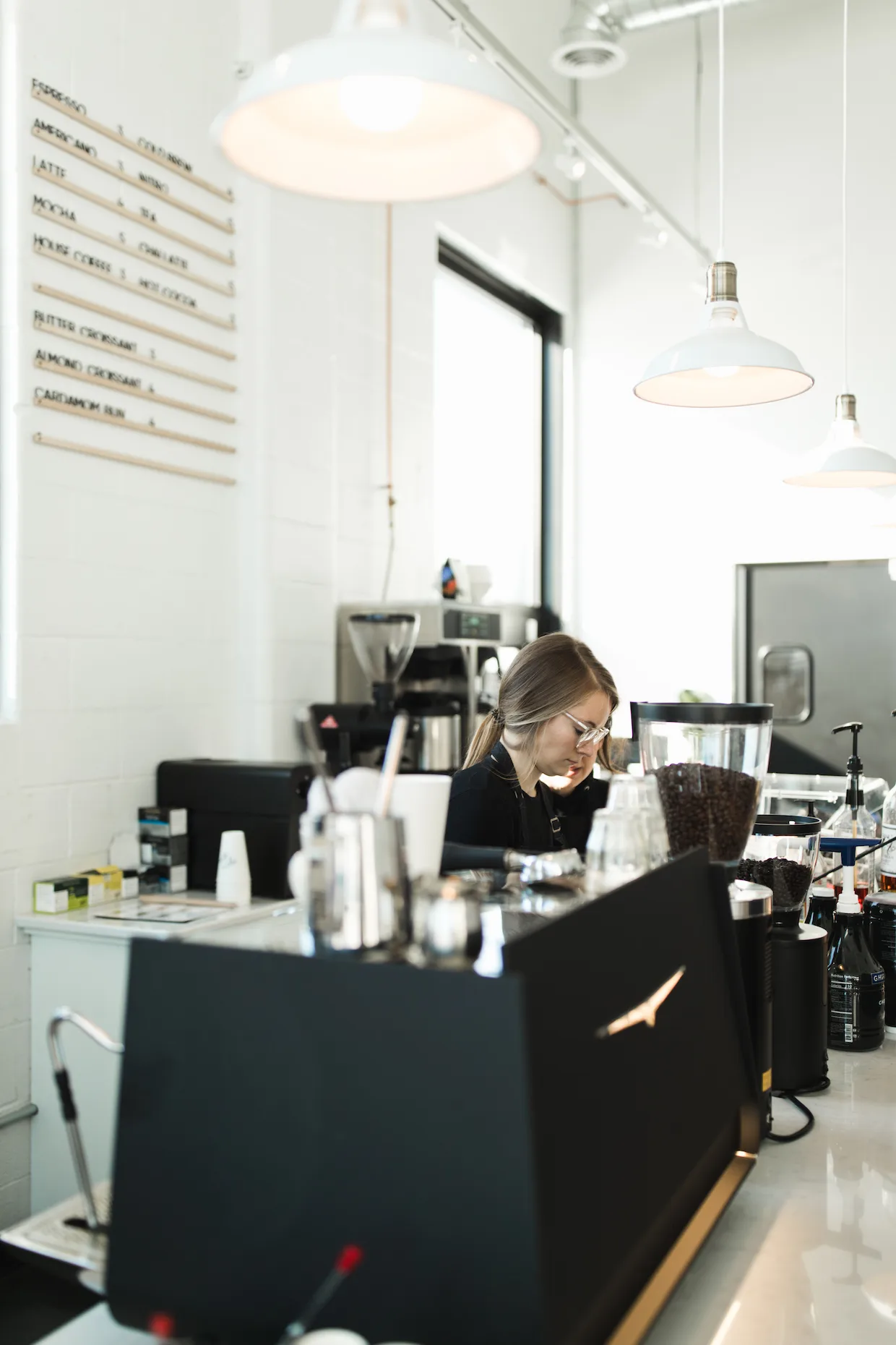 woman working at modern coffee shop counter with menu board and pendant lights., kinder coffee lab, saint cloud