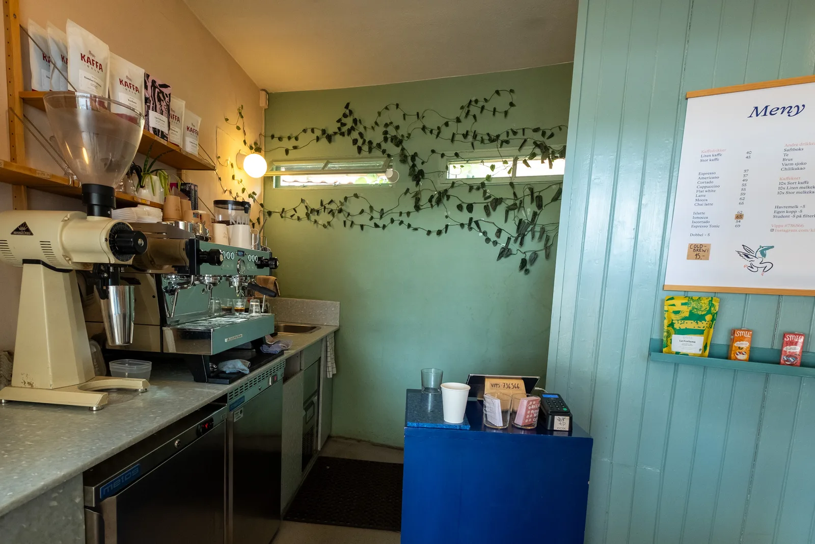 small coffee shop counter with espresso machine, grinder, and green botanical wall decor., kiosk! oslo, oslo