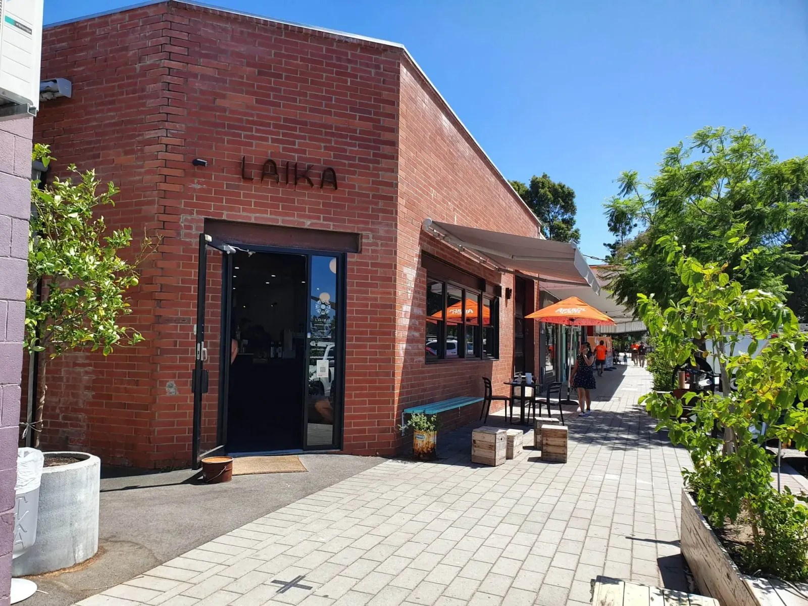 red brick building with "laika" signage, outdoor seating area with orange umbrellas., laika coffee, lathlain