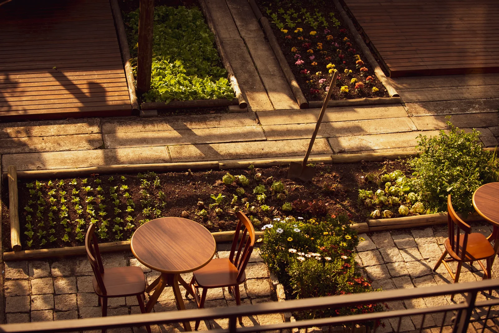 wooden tables and chairs surrounded by raised garden beds with flowering plants., leônidas bike café, curitiba