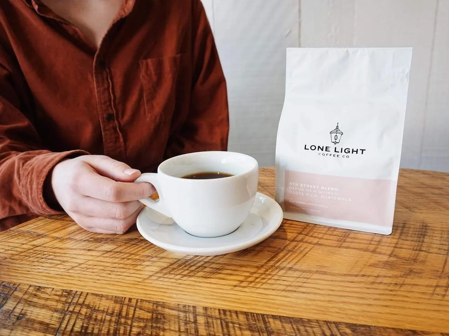 person in burgundy shirt holding white cup of coffee beside lone light coffee co. bag., lone light coffee, charlottesville