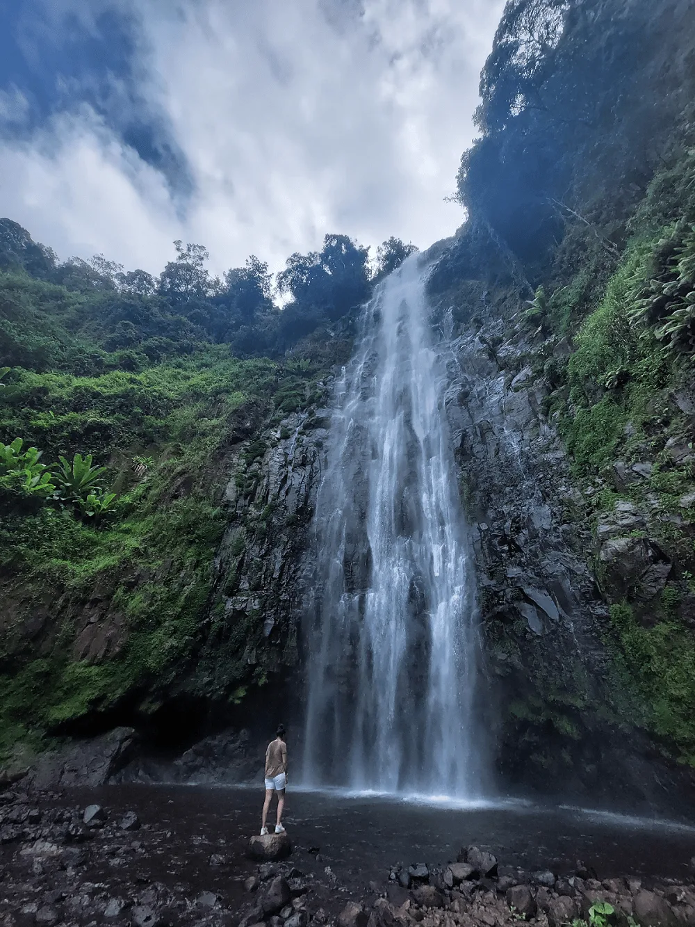 person standing before tall waterfall cascading down rocky cliff surrounded by lush green vegetation., materuni waterfalls an