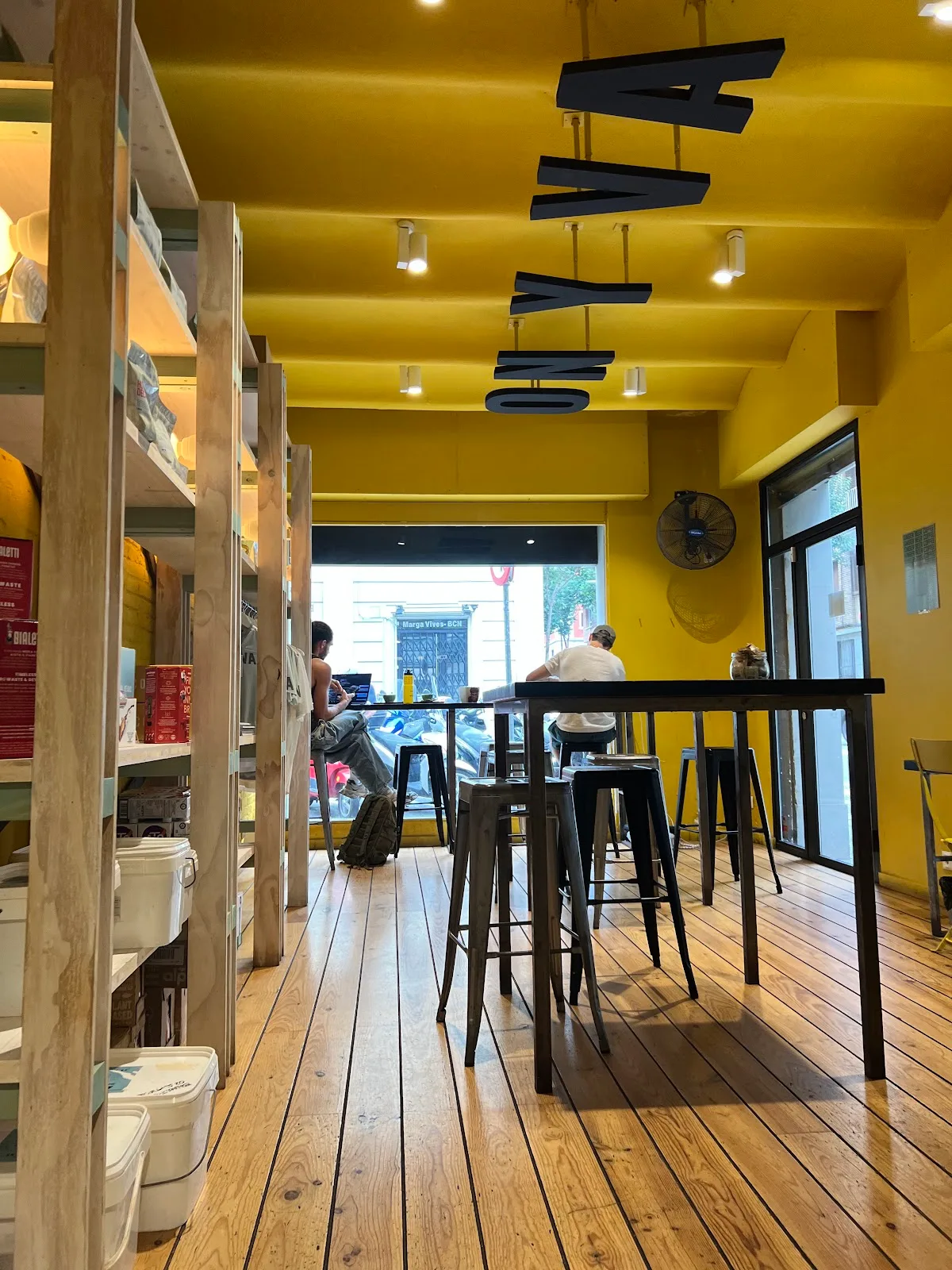 bright yellow cafe interior with wooden shelves, black bar seating, and customers., onyva coffee sant gervasi | cafe de espec